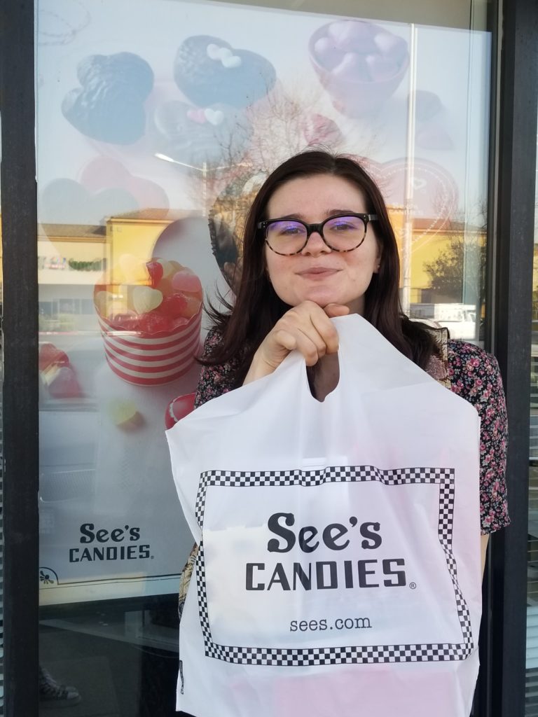 Young woman holding up a bag of See's candy outside of a See's candy store