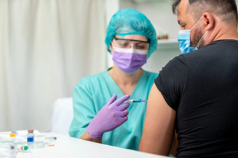 Mid adult man getting shot, nurse in protective workwear injecting vaccine